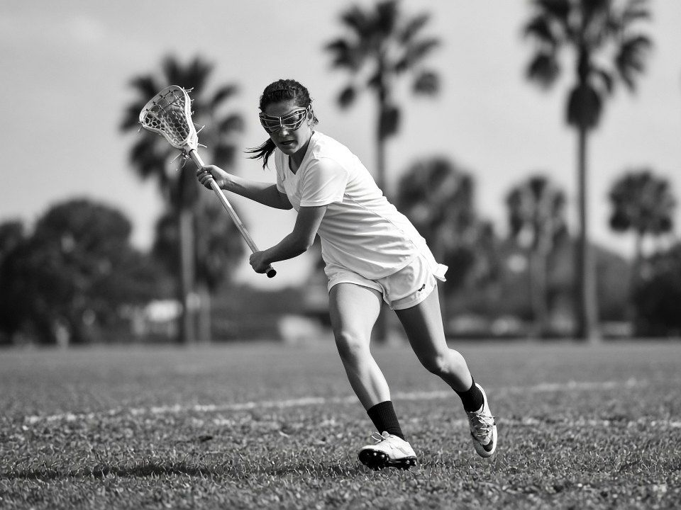 Black and white photo of a girls lacrosse player in goggles dodging with the ball, palm trees in the background.