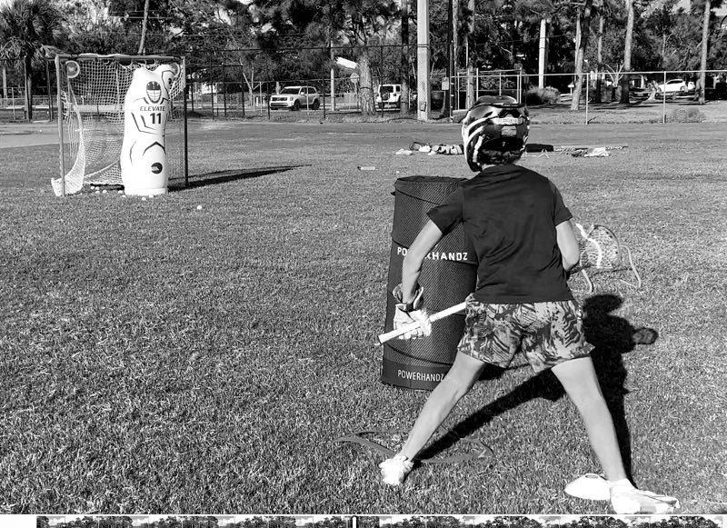 Athlete in a helmet practicing a lacrosse shot on a field near a goal.