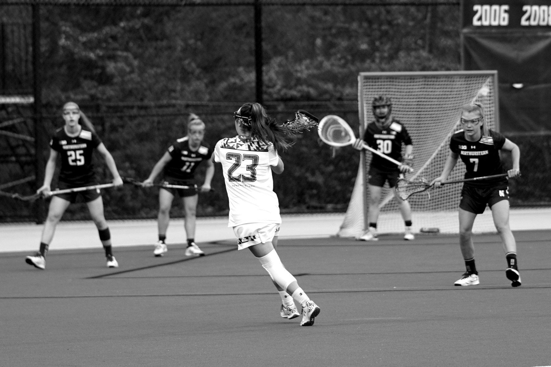 Four girls lacrosse players in uniform running a play on a turf field, in black and white.