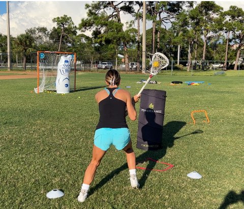 Athlete practicing a lacrosse shooting drill on a grass field.